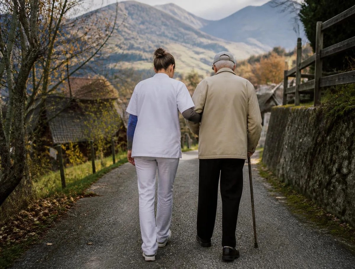 Betreuerin begleitet einen älteren Mann bei einem Spaziergang in einem Schweizer Dorf